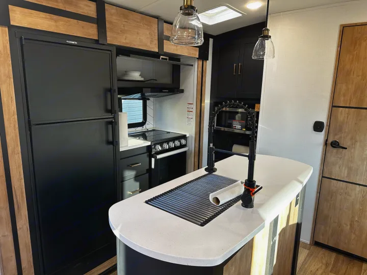 Inside a sleek RV rental, this kitchen boasts a Dometic fridge, black cabinetry, and an island with a sink and drying rack.. Keystone RV Outback Toy Hauler 2023