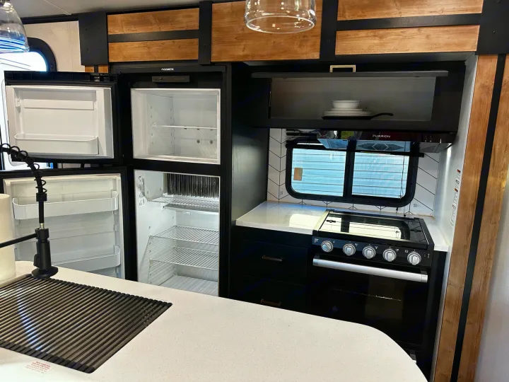 Clean, modern RV rental kitchen with an open double-door refrigerator, stove, and a white countertop with an integrated drainer.. Keystone RV Outback Toy Hauler 2023