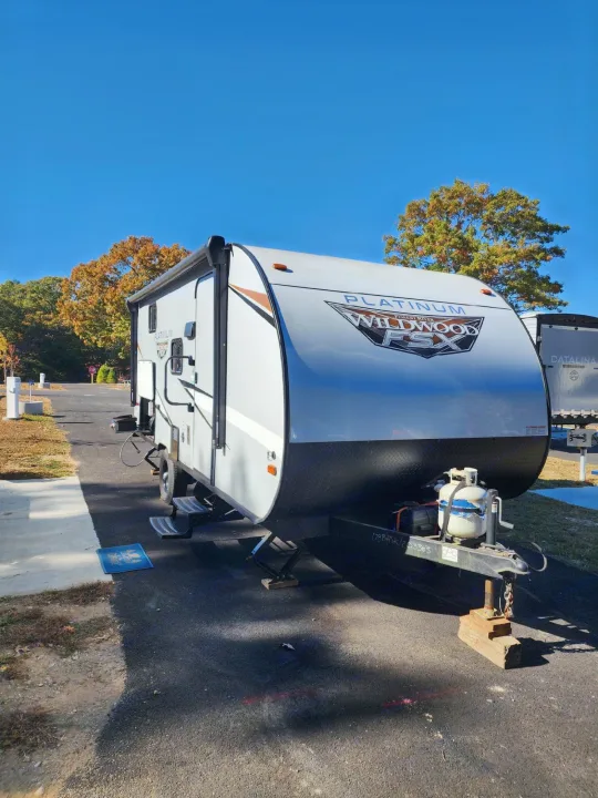 A white Platinum Wildwood travel trailer, an ideal RV rental, is parked on asphalt at a sunny campsite surrounded by autumn trees.. Forest River Wildwood FSX Plantium 2022