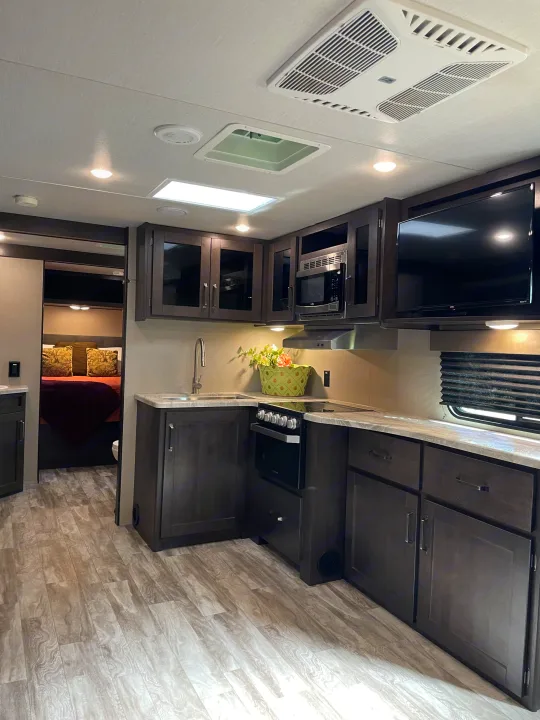 rv rental: Functional kitchen featuring dark wood cabinets, a sink, stove, microwave, TV, and a peek at the bedroom.. Grand Design Transcend 2021