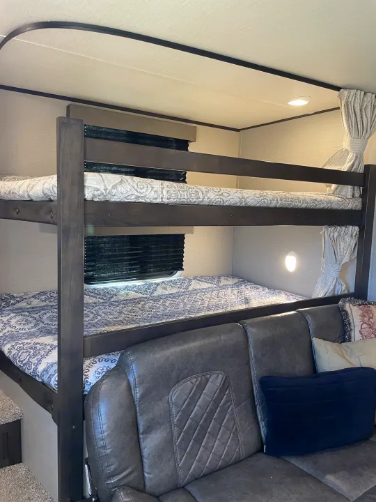 RV rental interior: cozy bunk beds with patterned bedding, a dark grey sofa, and ambient lighting in a camper.. Grand Design Transcend 2021
