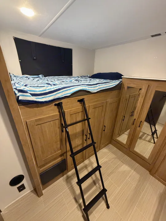 RV rental interior: a bunk bed with striped blanket, black ladder, and wood cabinets with mirrored doors, on tiled floor.. Coachmen Express 2024