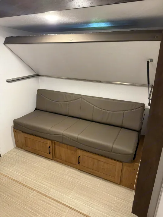 RV rental interior featuring a grey sofa, wooden cabinets below, and a raised bed platform above, ideal for compact living.. Coachmen Express 2024