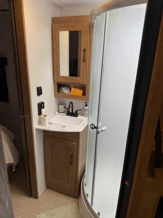 Inside an RV rental bathroom: wood vanity, white sink, black faucet, medicine cabinet, and a frosted glass shower enclosure.. Coachmen Express 2024