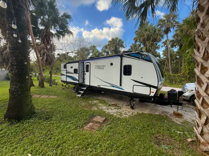 White Freedom Express RV rental parked amidst green grass and palm trees under a blue, cloudy sky. Perfect outdoor escape.. Coachmen Express 2024