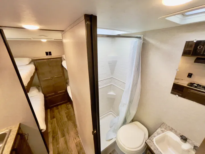 Inside an RV rental: view of bunk beds, bathroom (toilet, shower, sink), and kitchen reflection.. Sunset Park RV Sun Lite 2019