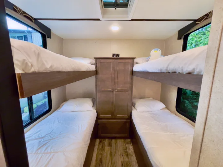 Interior of an RV rental: four bunk beds with white bedding, a central wardrobe, and windows looking out.. Sunset Park RV Sun Lite 2019