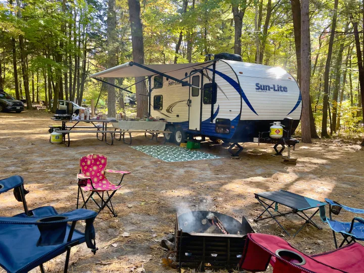 Enjoying a peaceful outdoor escape! This RV rental campsite features a Sun-Lite travel trailer, picnic area, and warm fire pit in a sunny forest setting.. Sunset Park RV Sun Lite 2019