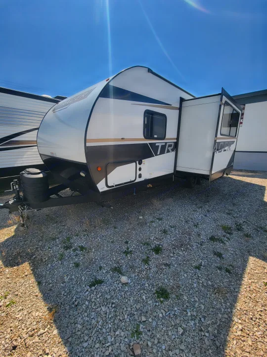 A modern white and black Tracer travel trailer, with an extended slide-out, parked on a gravel lot, available for RV rental.. Prime Time Tracer 2700BHSLE 2024