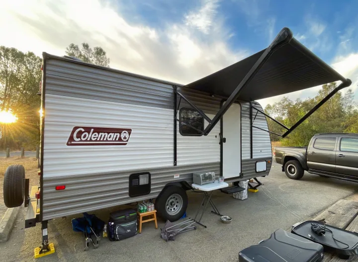 Cozy RV rental setup with a Coleman travel trailer, extended awning, and camping gear at sunset. Perfect outdoor adventure.. Keystone RV COLEMAN LANTERN LT 17B 2024