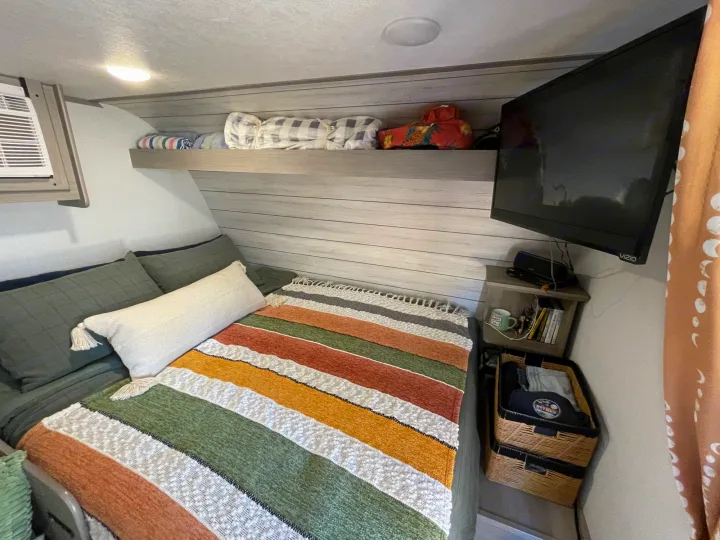 RV rental bedroom with a comfy bed, striped blanket, overhead shelf, TV, and storage baskets.. Keystone RV COLEMAN LANTERN LT 17B 2024