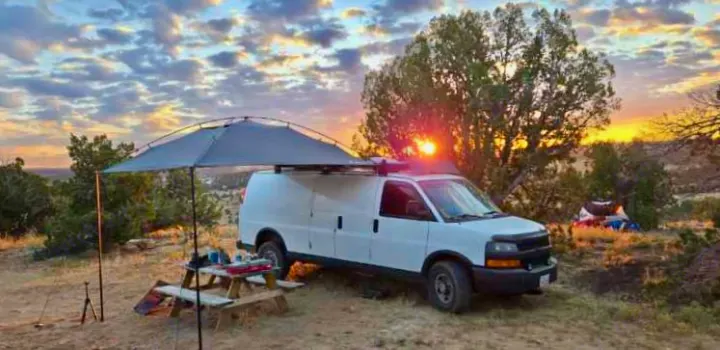 White camper van RV rental parked at a scenic campsite during sunset, with an extended awning & picnic table.. Chevrolet Express 3500 2012