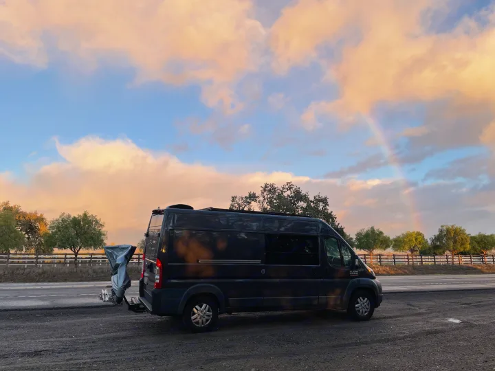 Stunning sunset sky with a rainbow over a black camper van RV rental, parked on a scenic roadside.. Ram Ram Promaster 2500 2018