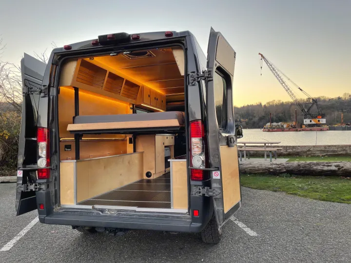 Explore this modern RV rental's rear, open doors showcasing a warm, lit wood interior with a raised bed & storage. Parked by water.. Ram Ram Promaster 2500 2018