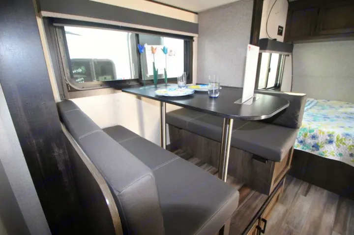 This RV rental offers a compact dinette with gray seating & a window, next to a bed with a floral quilt, ideal for travel.. Starcraft Autumn Ridge 180BHS 2023