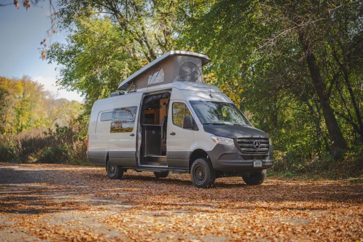 Explore nature with this Mercedes Sprinter camper van, an ideal RV rental featuring a pop-top roof, parked on a leafy path in autumn woods.. Mercedes Sprinter 2023