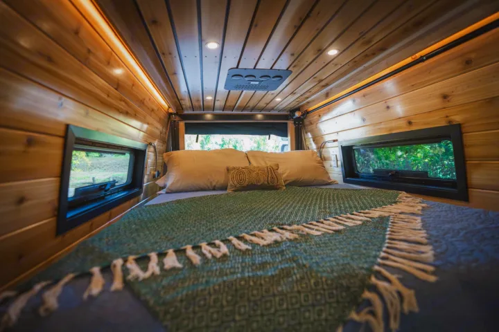 Inside a comfortable RV rental bedroom, featuring warm wood walls, ceiling lights, and scenic windows.. Mercedes Sprinter 2023