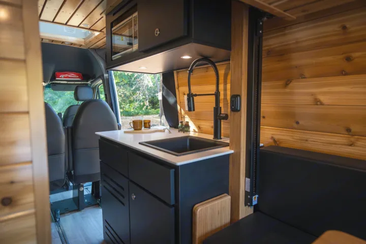 Cozy RV rental interior featuring a modern kitchen with black cabinets, white counter, sink, and microwave, surrounded by warm wood paneling.. Mercedes Sprinter 2023