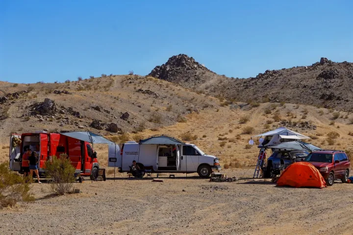 Overland RV rental setup in the desert, featuring camper vans, a rooftop tent on an SUV, and an orange ground tent.. Chevrolet Express 3500 2012