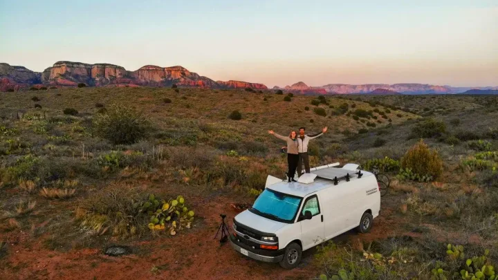 Couple celebrating on their RV rental van, surrounded by desert shrubs and red rock mountains at sunset. Van life adventure!. Chevrolet Express 3500 2012