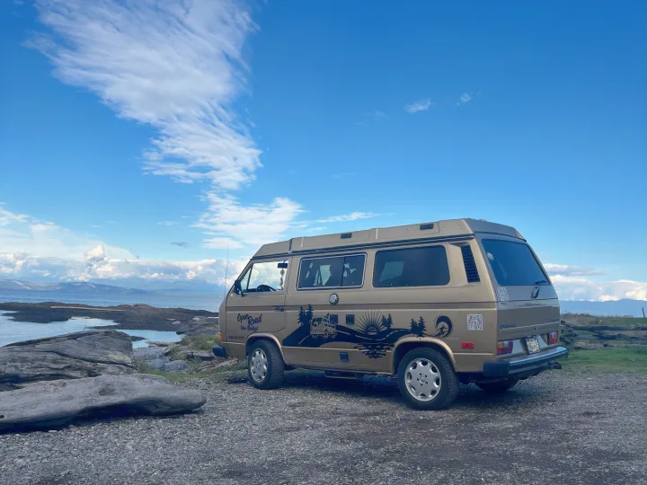 Tan RV rental van parked by the coast, overlooking water, mountains & a blue sky. Ready for open-road adventures!. Volkswagen T3 Westfalia 1985