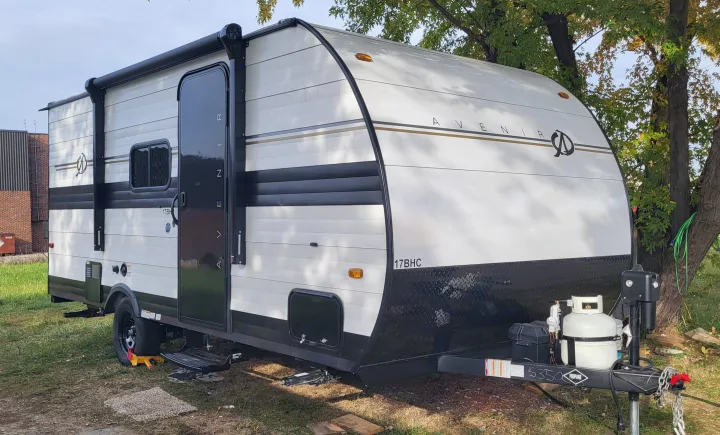 A white and black Avenir 17BHC travel trailer, ideal for an RV rental, parked on grass near trees.. A Liner Ascape 2024