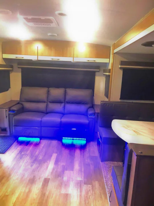 Cozy RV rental interior with a modern reclining sofa and dining area. Blue under-lighting adds ambiance to the wood-style floor.. Grand Design Imagine 2023