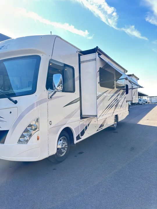 Large white RV rental with slide-out and awning deployed, parked on an asphalt lot under a blue sky.. Thor A.C.E. 2018