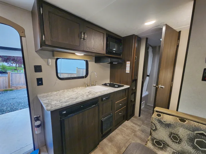 Interior of an RV rental featuring a compact kitchen with sink, stovetop, microwave, and fridge, adjacent to a seating area.. Keystone RV Springdale 2018
