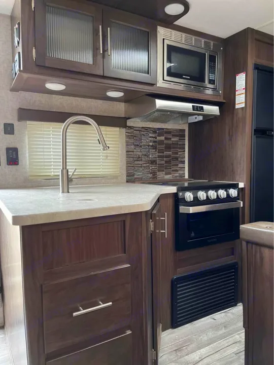 Modern RV rental kitchen with dark wood cabinetry, stainless steel sink, microwave, and stove/oven combo. Ready for your adventure!. Forest River Cherokee 2018