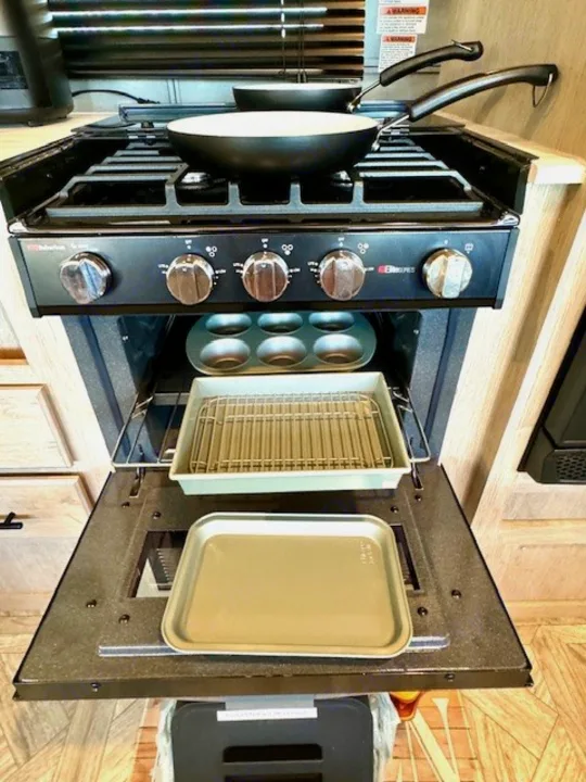 Inside an **rv rental** kitchen: a gas stove with pans, plus an open oven displaying a muffin tin and baking pans.. Forest River Wildwood 2024