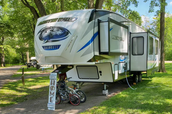 White fifth-wheel RV, a potential RV rental, sits ready for camping at a shaded site with bikes and a 'Happy Home Campers' sign.. Forest River Cherokee Arctic Wolf 2019