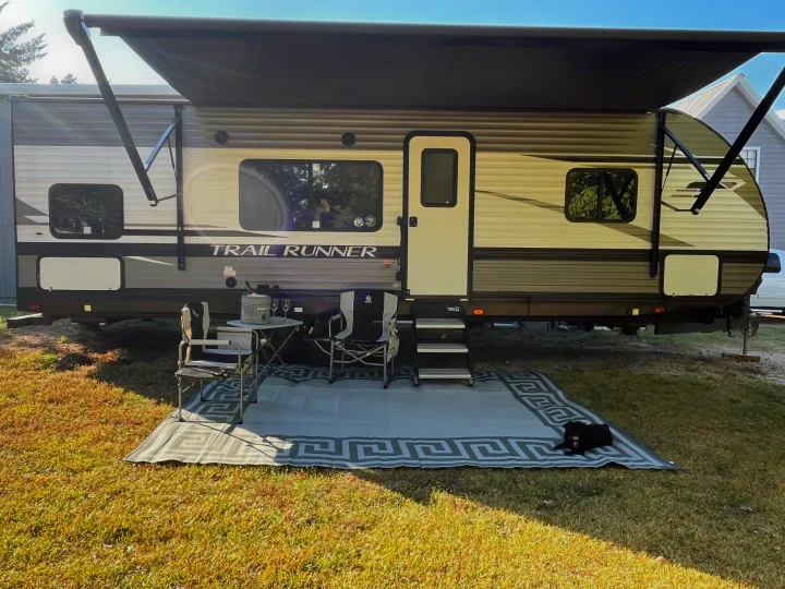 A sunny RV rental campsite featuring an extended awning, outdoor seating, and a black dog relaxing on a mat.. Heartland RVs Trail Runner 2022