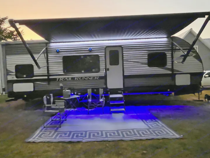 An RV rental at dusk, awning extended with bright white lights, and blue under-glow. An outdoor rug, chairs, and table are set up.. Heartland RVs Trail Runner 2022