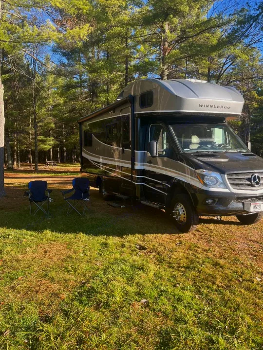A black Winnebago RV rental awaits adventure in a sunny, wooded campsite with two blue chairs on the grass.. Winnebago View 2019