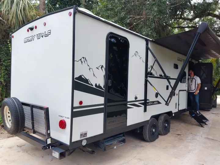This Grey Wolf RV rental, with its extended awning, rear rack, and steps, offers outdoor adventure. A man stands by the door.. Forest River Cherokee Grey Wolf Black Label 2021