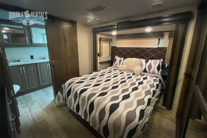 Step inside this cozy RV rental's master bedroom, complete with a modern bed and an en-suite bathroom visible through a barn door.. Keystone Montana 2019