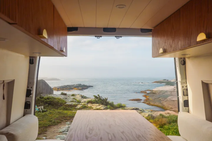 Looking out from an RV rental: a serene coastal view with rocky shores. Inside, wooden cabinets & table are visible.. Mercedes-Benz Sprinter 2020