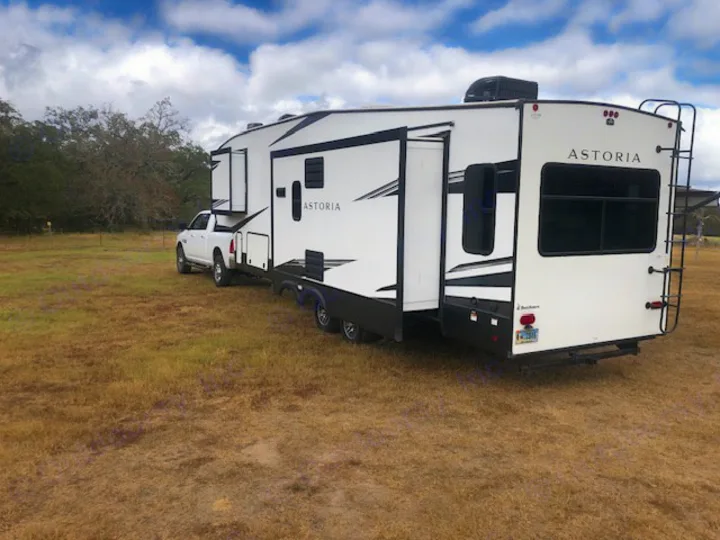 White pickup truck towing a large Astoria RV rental, a travel trailer parked in a dry, grassy field with slide-outs extended.. Astoria 1500 2021