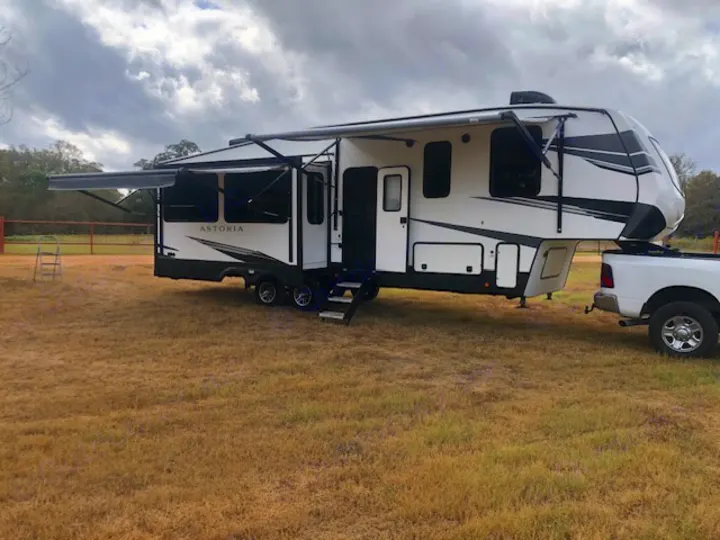 This white fifth-wheel RV rental, with awning extended, is ready for your next adventure, hitched to a pickup truck on dry grass.. Astoria 1500 2021