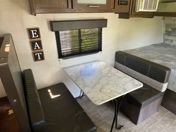 This RV rental shows a cozy dinette with dark benches, a marble table, and a bed beyond, perfect for compact living.. Dutchmen Coleman Lantern 2021