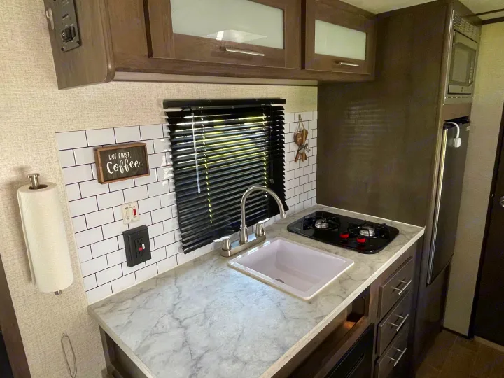 Compact RV rental kitchen with sink, two-burner stove, microwave, refrigerator, and marble-look countertop.. Dutchmen Coleman Lantern 2021