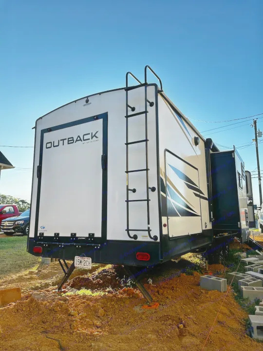 This Outback RV rental, a white travel trailer with rear ladder & side slide-out, is parked on dirt with stabilizers deployed.. Keystone RV Outback Toy Hauler 2023