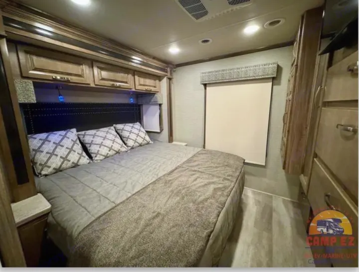 Modern RV rental bedroom with a large bed, dark headboard, overhead cabinets, blue accent lights, and light wood flooring.. Jayco Seneca 2020