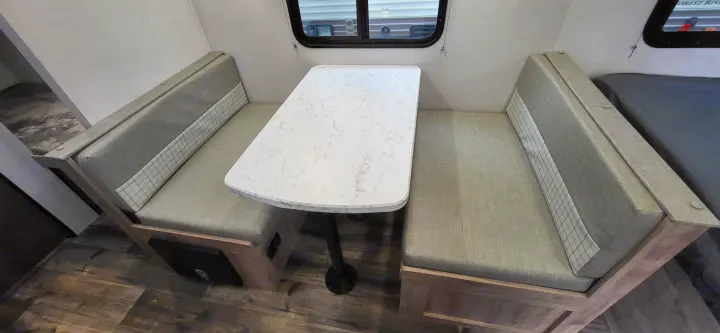 Bright RV rental interior: a dinette with grey cushioned benches and a white marble-look table, beside a window and bed area.. Gulf Stream Ameri-Lite 2024