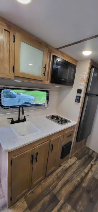 Inside an RV rental: a bright, compact kitchen area featuring a sink, two-burner stove, microwave, and wooden cabinets.. Gulf Stream Ameri-Lite 2024