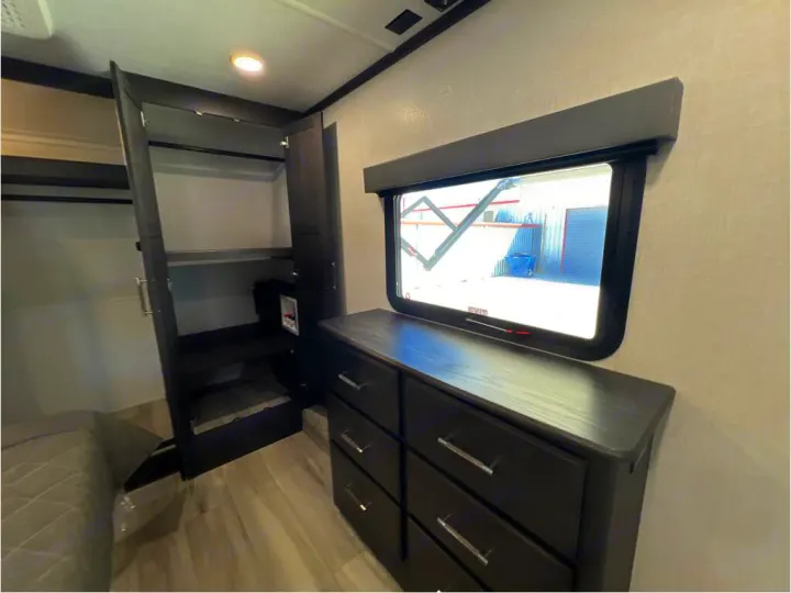 RV rental bedroom: dark wood wardrobe with shelves & washer/dryer hookup. Features a dresser, window, and bed.. Keystone Alpine 2025