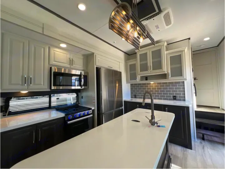 Modern RV rental kitchen with white cabinets, stainless appliances, large island, gas range, and gray tile backsplash.. Keystone Alpine 2025