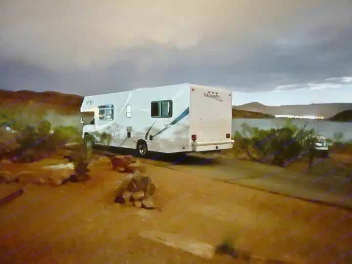 Nighttime view of a Majestic RV rental, a Class C motorhome, parked on a dirt road beside a large body of water with distant lights.. Thor Majestic 2013