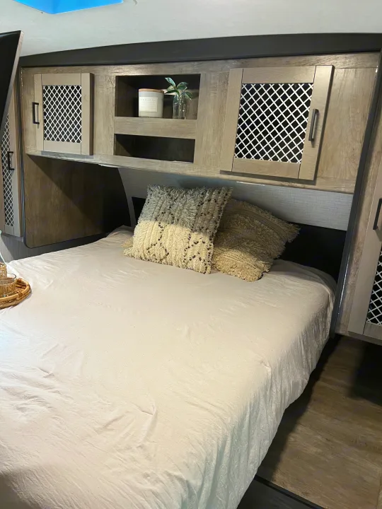 This RV rental bedroom features a cozy bed with white linens and patterned pillows, overhead cabinets, and built-in shelving.. Heartland RVs North Trail 2021